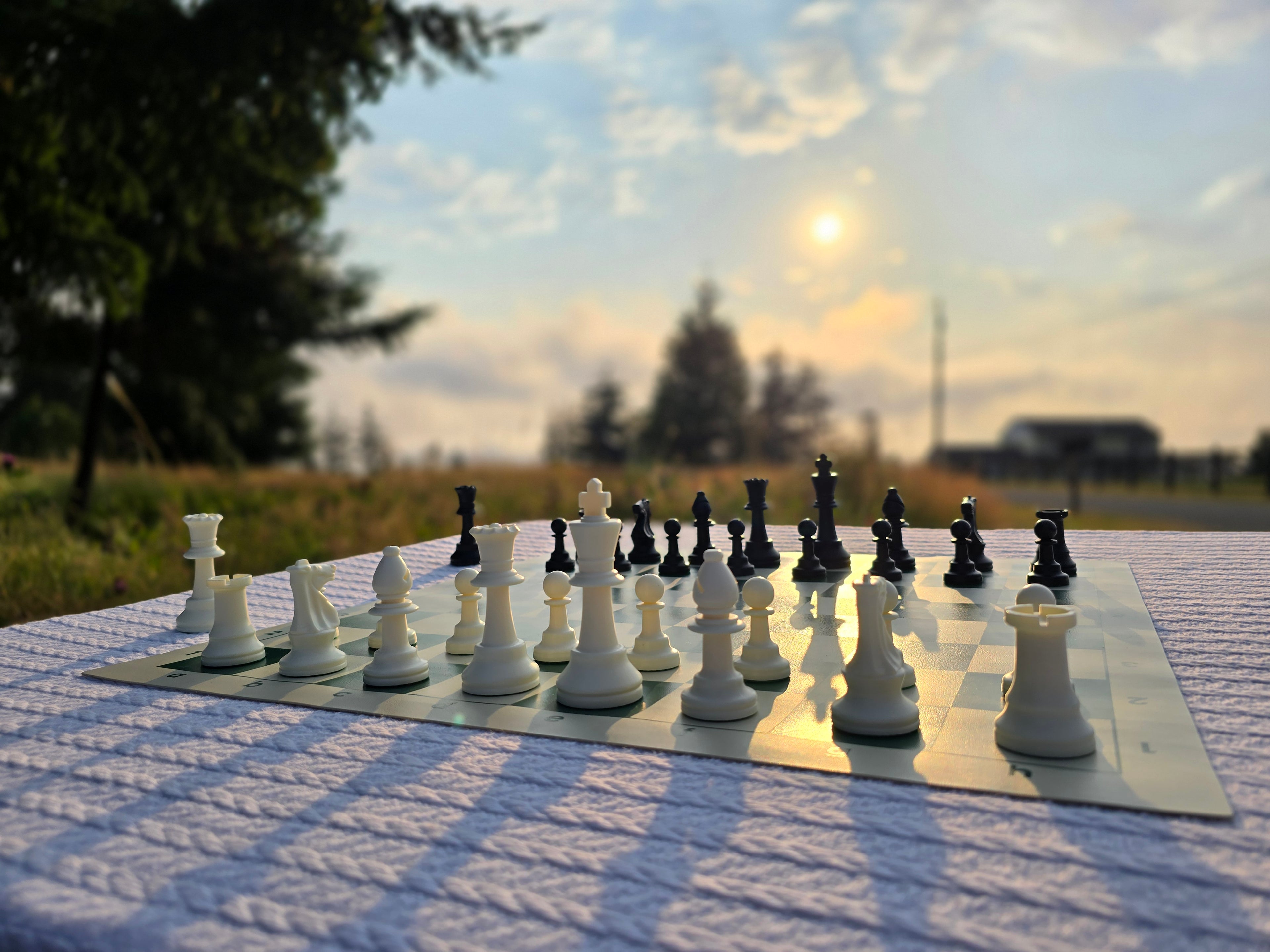 Tournament Chess Set on a Vinyl Chess Board on a White, Knitted Tablecloth in a Meadow with a Setting Sun, and Blurred Background with a Farmhouse in the Distance