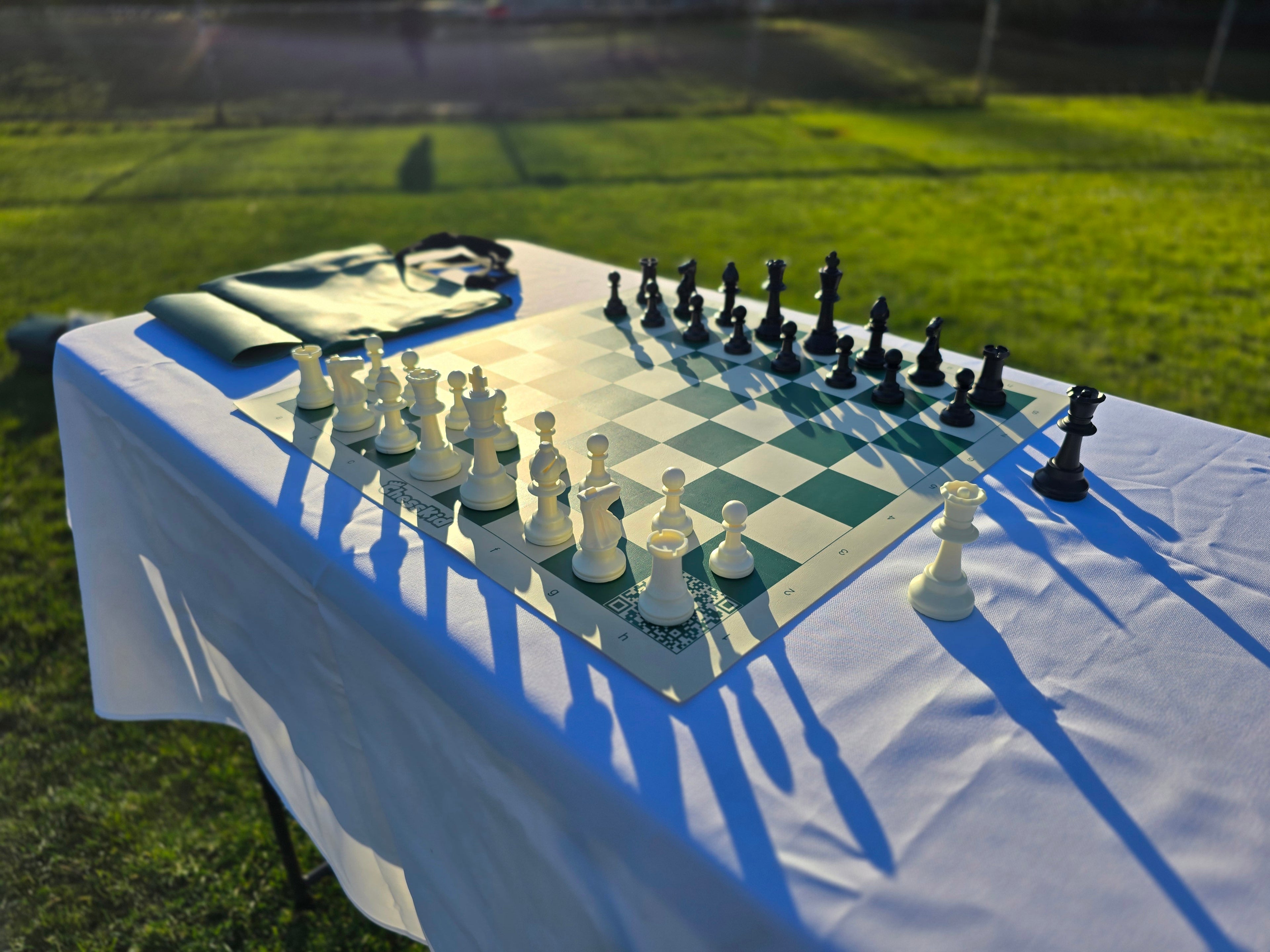ChessKid Tote Chess Set with Vinyl Chess Board on White Cloth Table in Park (Under Shining Sun, Angled Shot from H1)