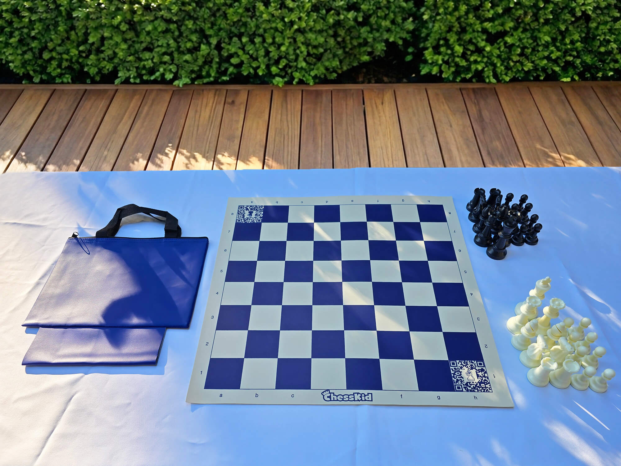 A ChessKid blue vinyl roll-up chess board set with a matching blue carrying tote bag. The set includes a full collection of black and white plastic chess pieces arranged on a white outdoor table.