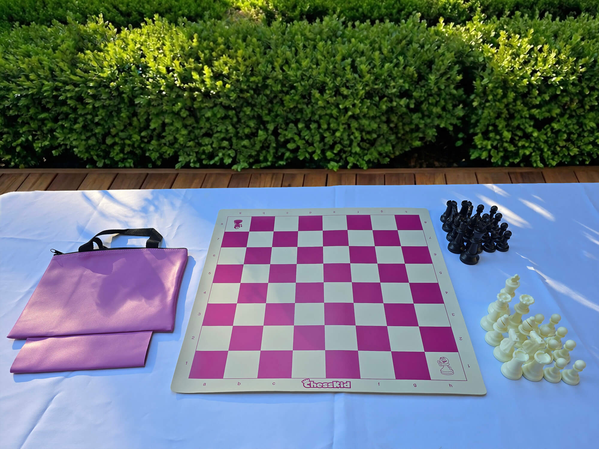 A ChessKid pink vinyl roll-up chess board set with a matching carrying tote bag. The set includes a full collection of black and white plastic chess pieces arranged on a white outdoor table.