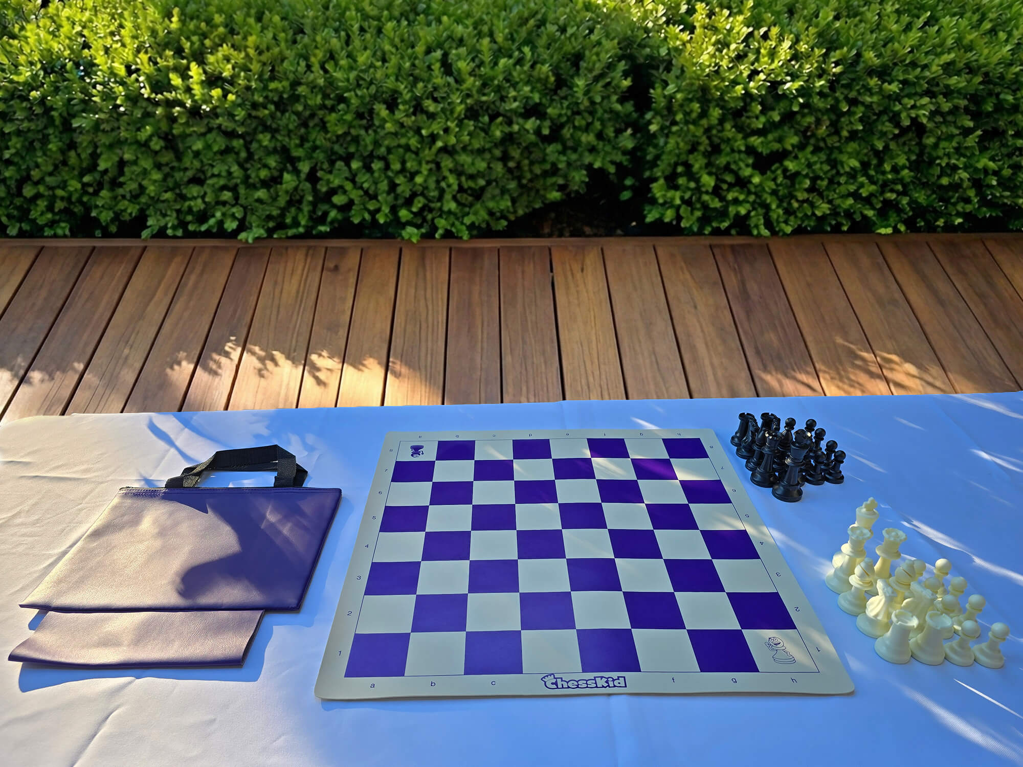 A ChessKid purple vinyl roll-up chess board set with a matching carrying tote bag. The set includes a full collection of black and white plastic chess pieces arranged on a white outdoor table.