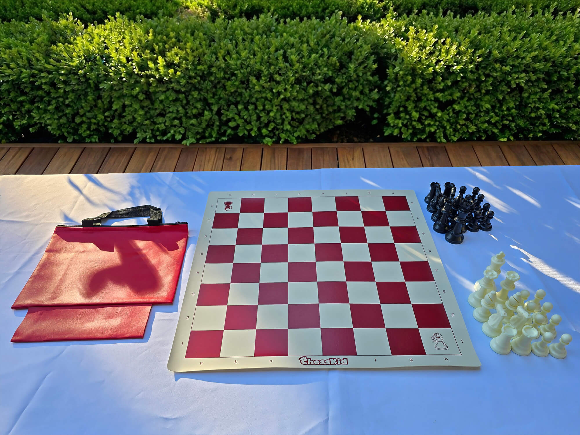A ChessKid red vinyl roll-up chess board set with a matching carrying tote bag. The set includes a full collection of black and white plastic chess pieces arranged on a white outdoor table.