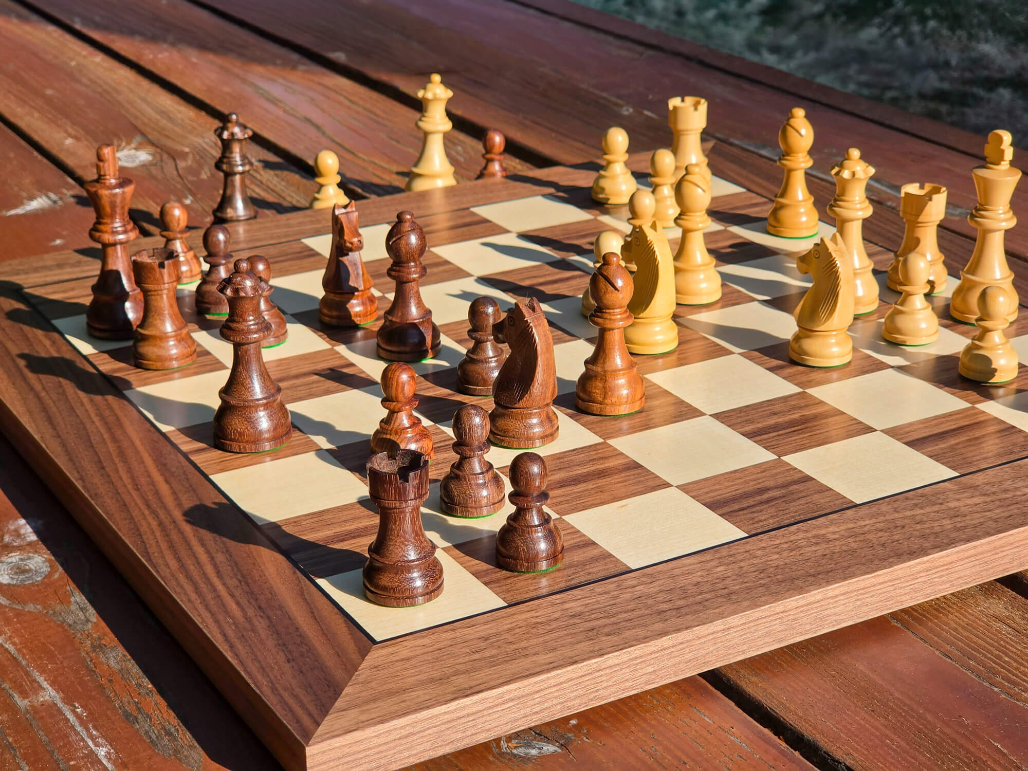 Elevated corner shot of the DGT Timeless chess pieces set up on a wooden board resting on a park bench with a nature background.