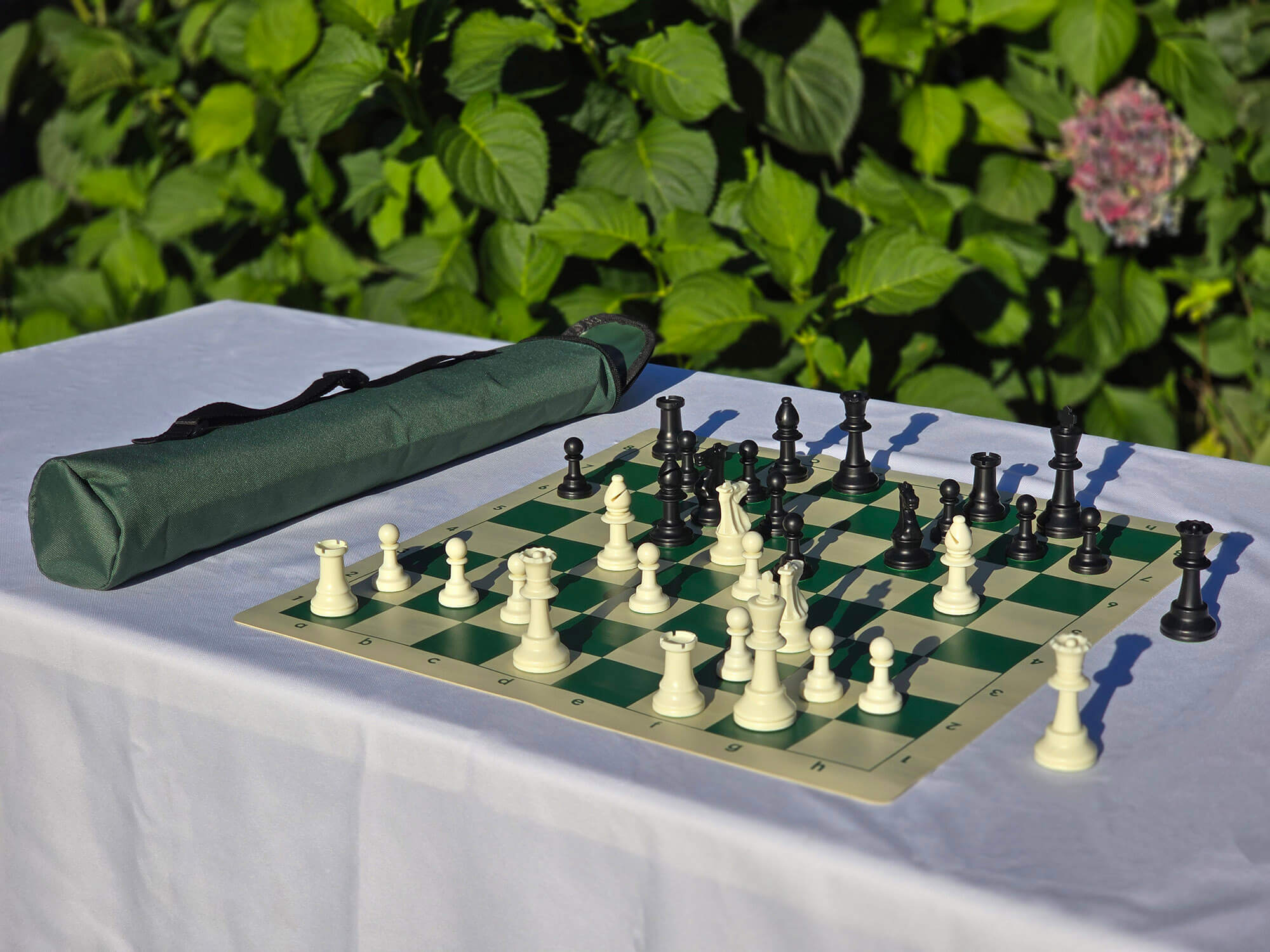 Tournament weighted chess set with green roll-up vinyl board and matching green quiver-style carrying bag with shoulder strap, displayed outdoors on a white table.
