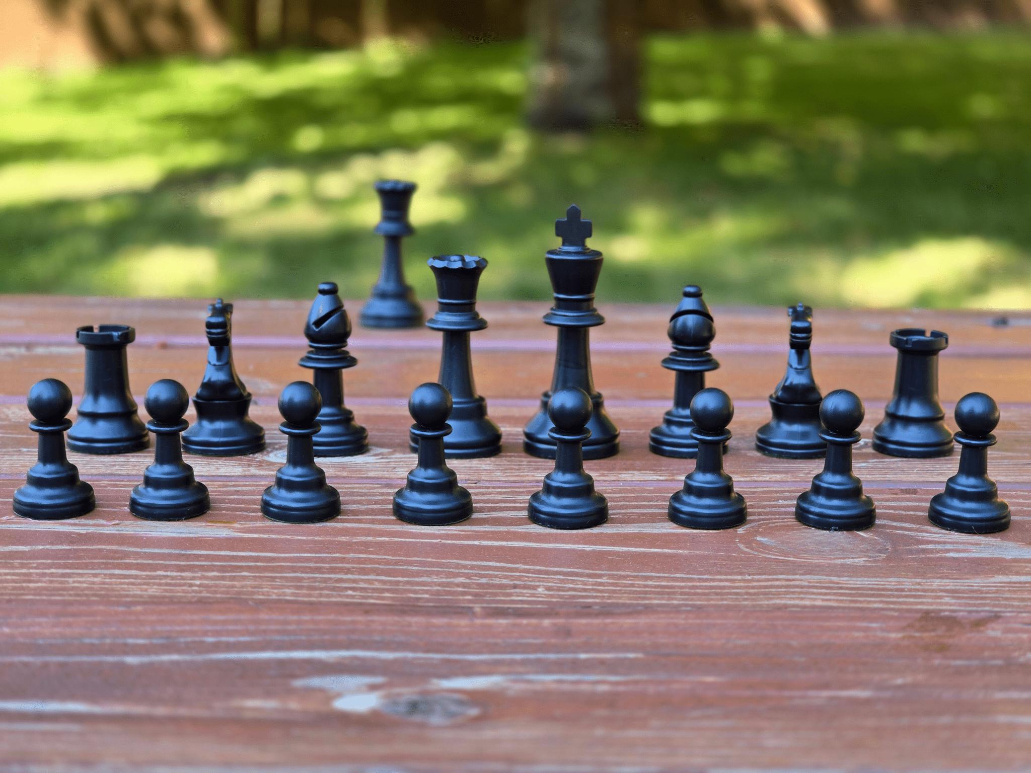 Black Plastic Chess Pieces in Standard Order on a Dark Brown Park Bench Against a Blurred Lush Green Background