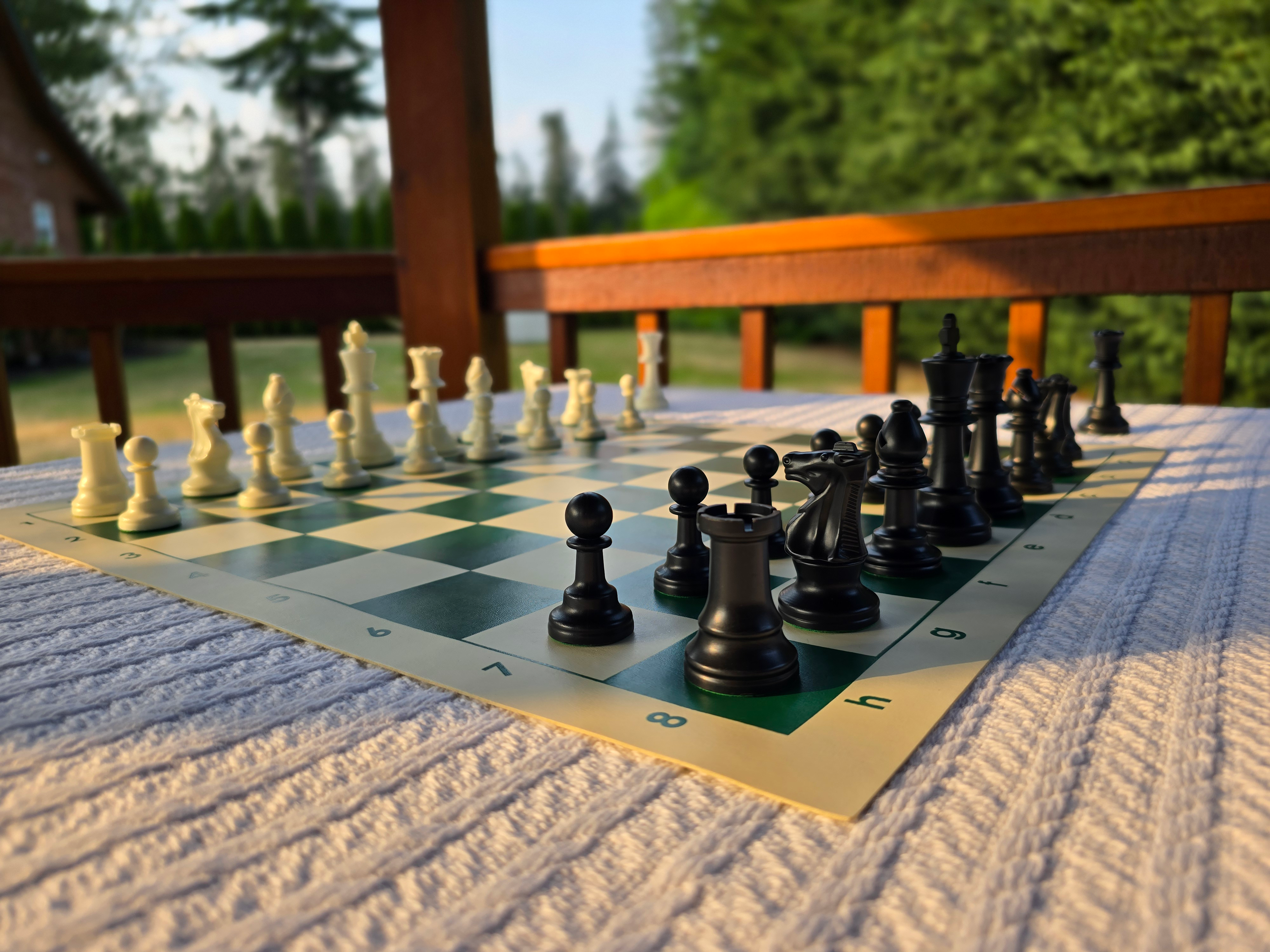 Tournament Chess Set on a Vinyl Chess Board on a White Knit Tablecloth in a Gazebo Against a Lush Green Background