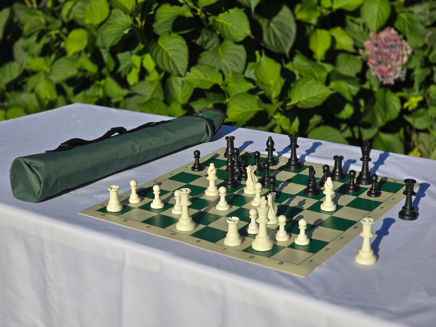 Tournament Chess Set on a Vinyl Chess Board with a Quiver Carrying Case on a White Cloth Table Against a Rose Bush Background (Angled Shot)