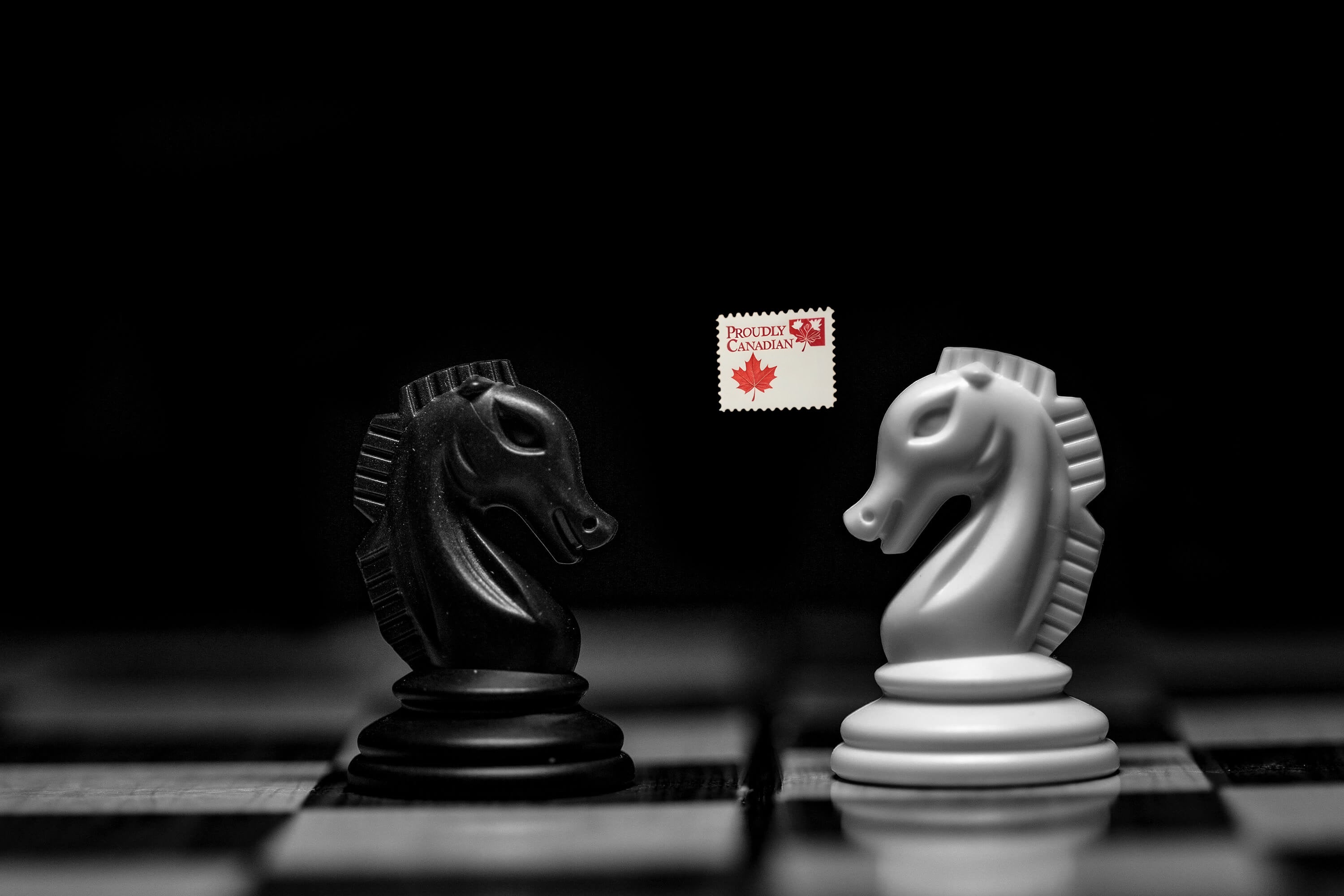 A Black and White Knight Facing Each Other Down on a Chess Board with a "Proudly Canadian" Stamp in the Background