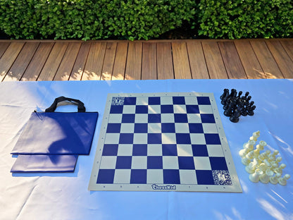 A ChessKid blue vinyl roll-up chess board set with a matching blue carrying tote bag. The set includes a full collection of black and white plastic chess pieces arranged on a white outdoor table.