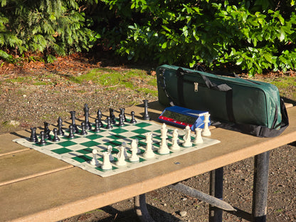 Chess set with pieces on a board outdoors with DGT North American Clock, surrounded by greenery.