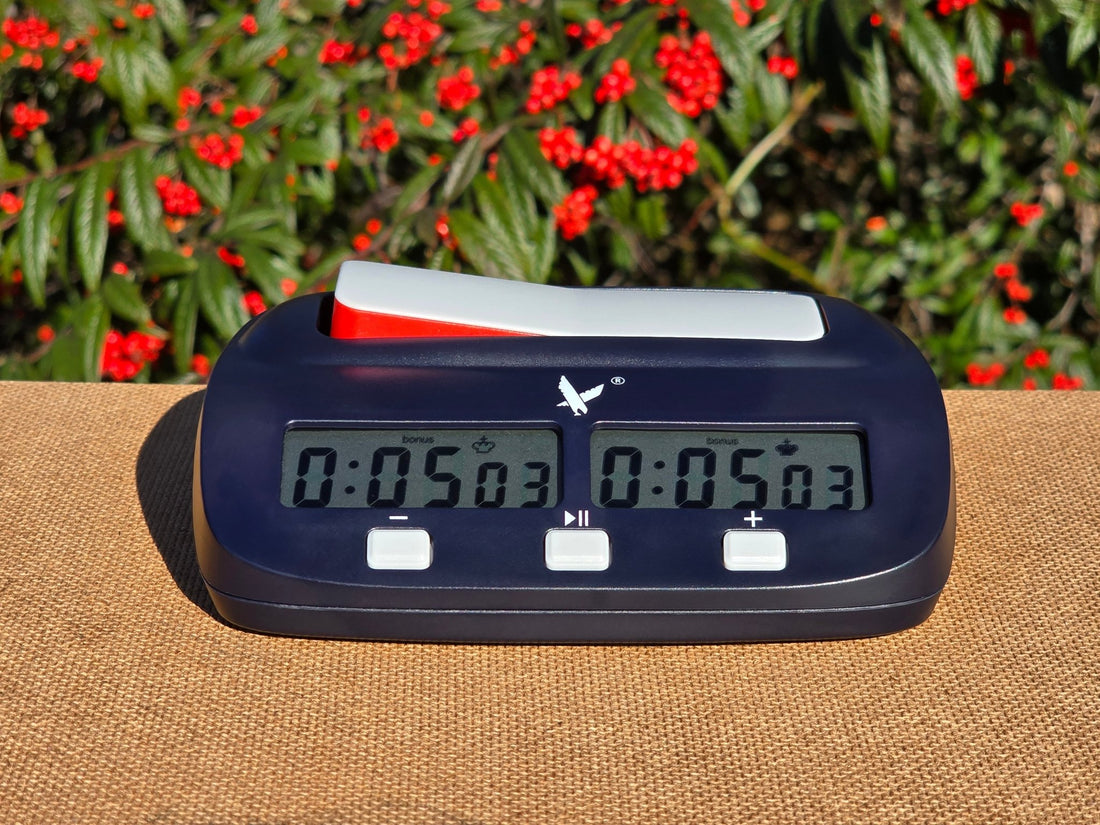 A navy blue Leap KK9908 digital chess clock sitting on a textured surface with a background of green leaves and red berries.