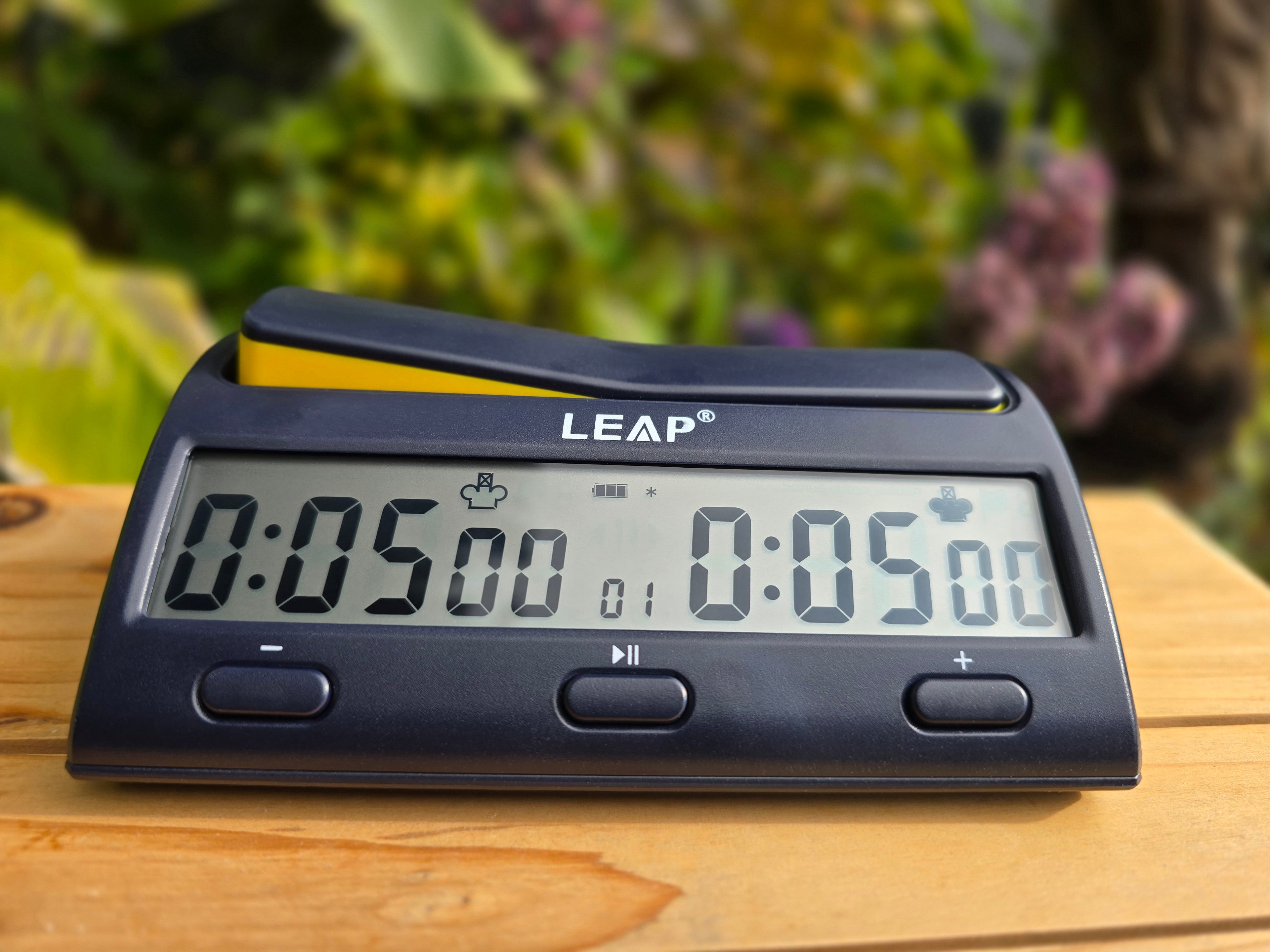 Leap KK9909 Chess Clock on Wooden Board with pink flowers and bush in background.