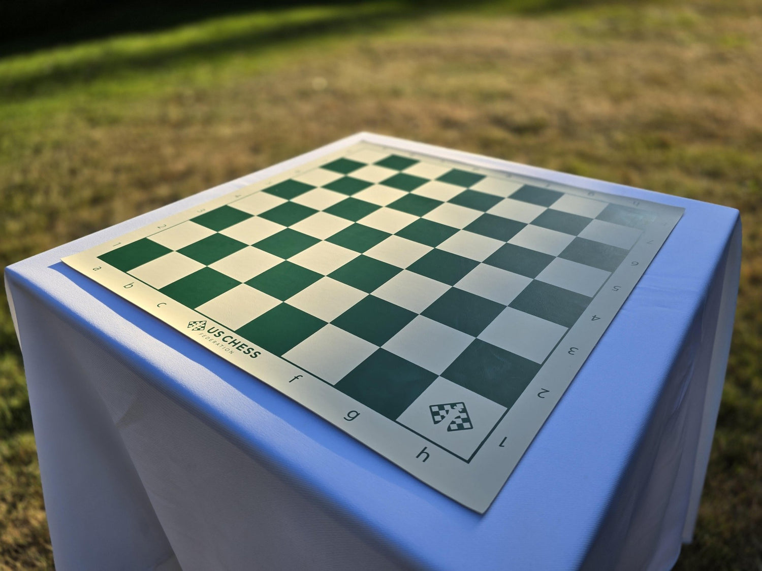 US Chess Federation Vinyl Chess Board on a White Cloth Table in a Sunlit Grass Field (Angled H1 Perspective)