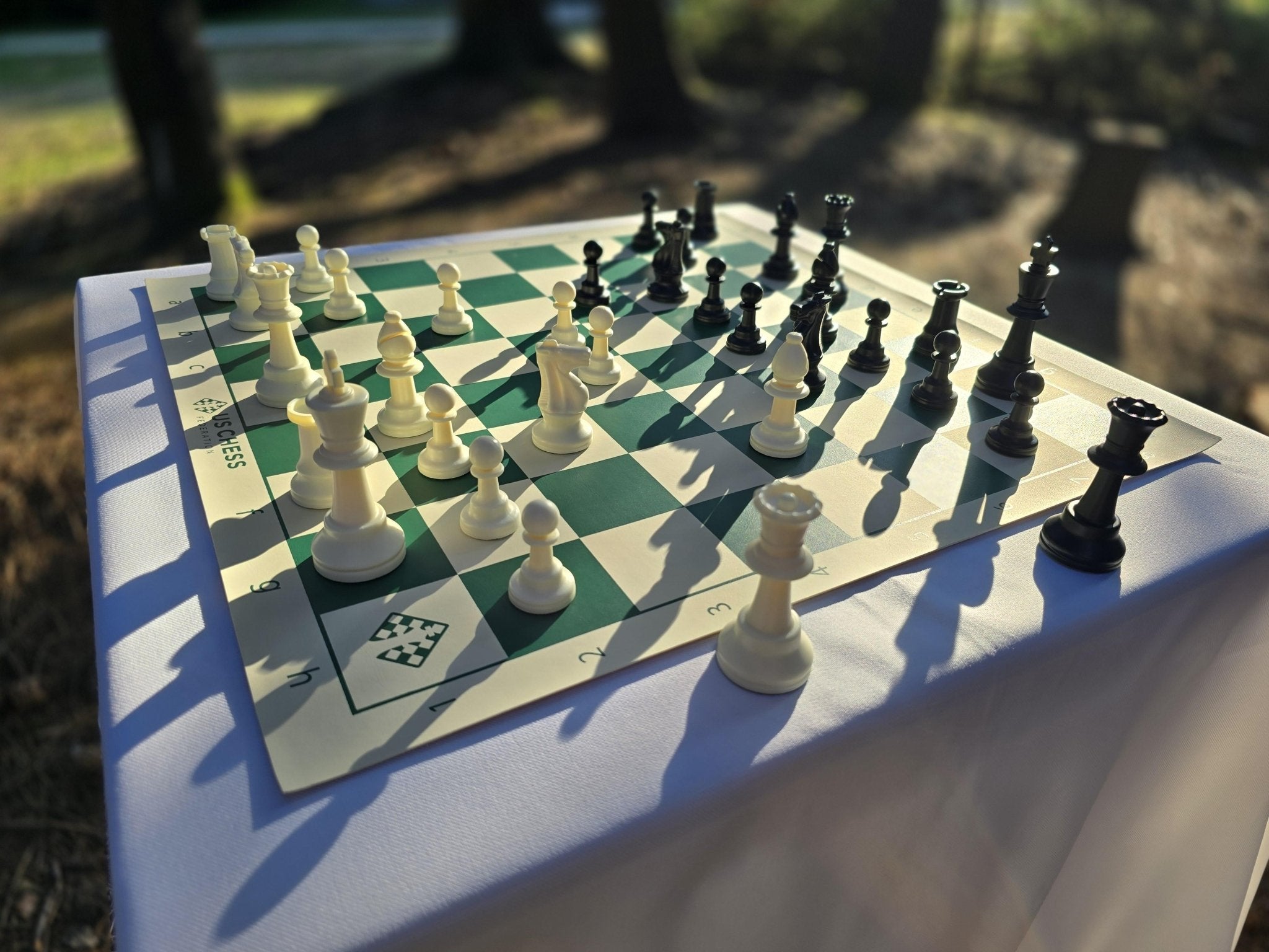 US Chess Federation Tournament Chess Set with USCF Vinyl Chess Board on a White Cloth Table in a Park at Twilight with Sunlight Glinting Off It