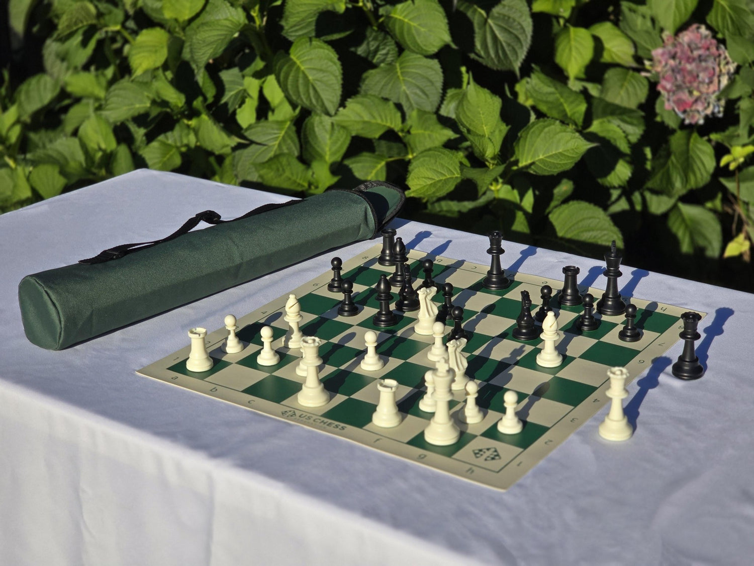 US Chess Federation Tournament Chess Set with Vinyl Chess Board and Quiver Carrying Bag on a White Cloth Table Against a Rose Bush Background (Angled Shot)