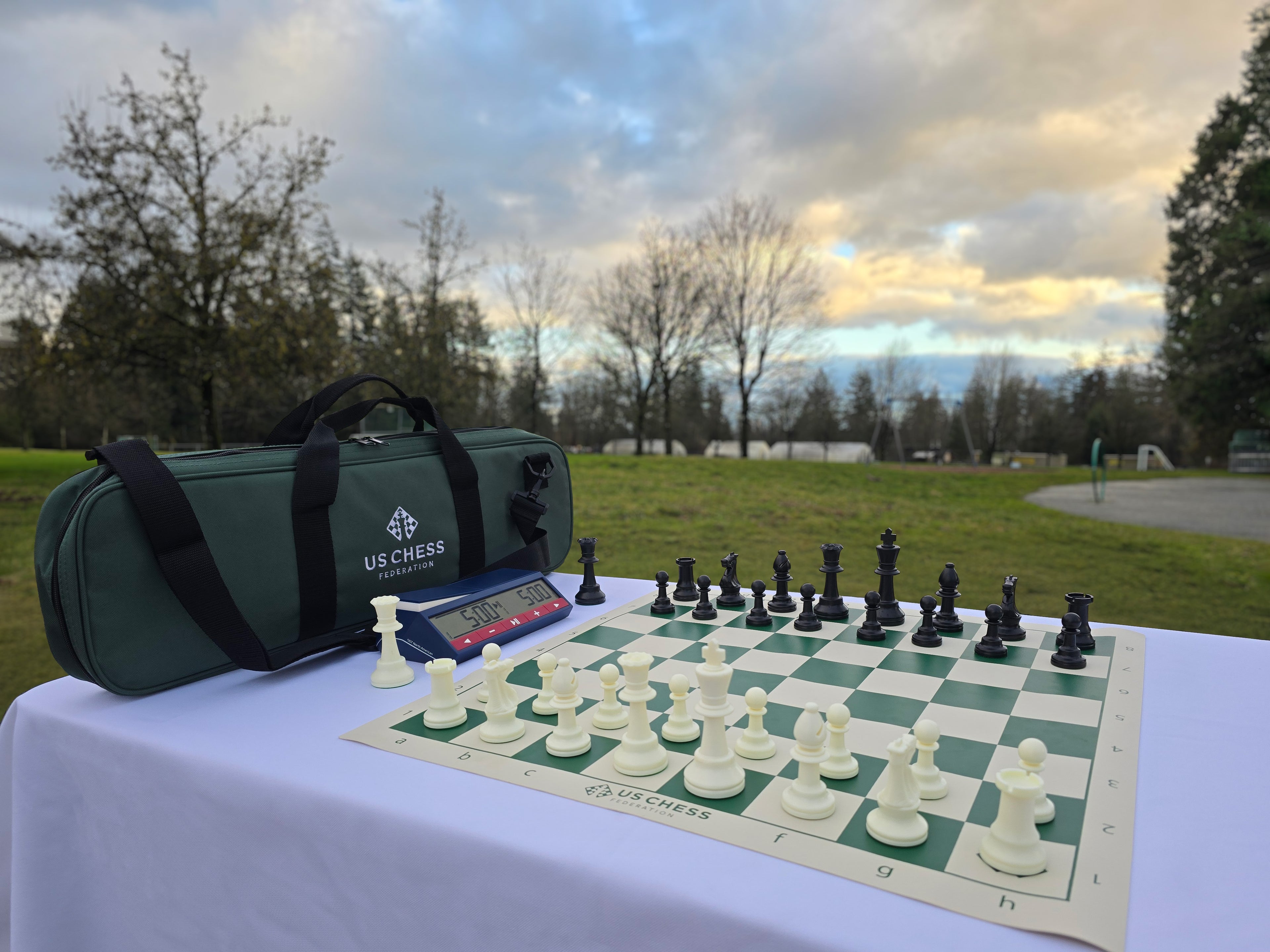USCF Deluxe Chess Set with Vinyl Chess Board and DGT North American Chess Clock 2024 in Park Against a Beautiful, Golden Cloud and Blue Sky Background