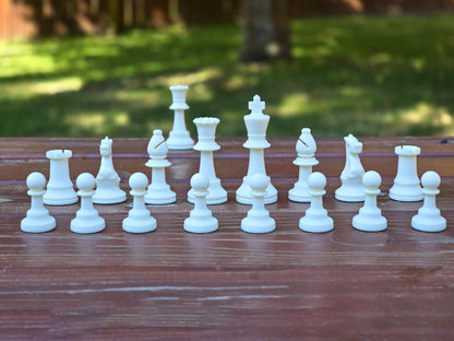 White Plastic Chess Pieces in Standard Order on a Dark Brown Park Bench Against a Blurred Lush Green Background