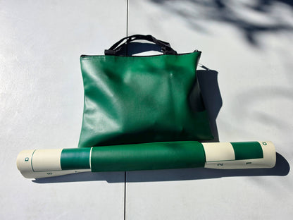 Packed green vinyl tote bag holding chess pieces with the rolled-up vinyl board secured on the exterior bottom loop.