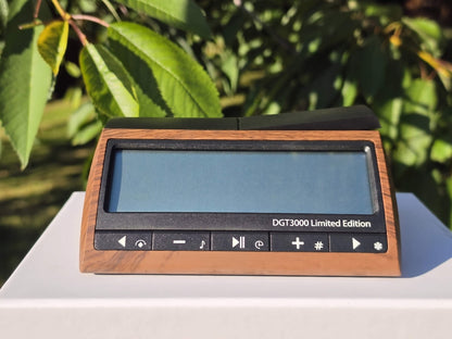 Front view of the DGT 3000 Limited Edition digital chess clock with a wood-grain finish, displayed outdoors against a natural green foliage background.