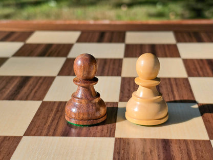 Close-up of dark brown and natural wood Pawn chess pieces from the DGT Timeless set resting on a wooden board with a nature background.