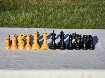 Complete set of black and natural wood Judit Polgar chess pieces lined up outdoors on a white tablecloth.