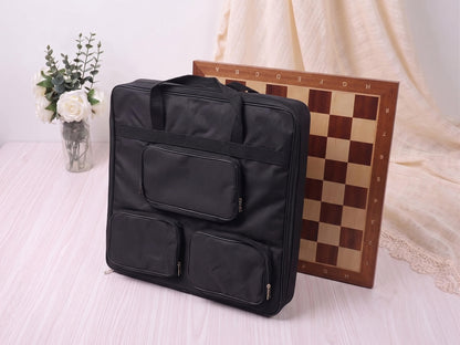 A large black padded nylon carrying bag for a Monarch series wooden chess board, featuring three external zippered accessory pouches, sitting in a studio setting with a large wooden chessboard and a vase of flowers.