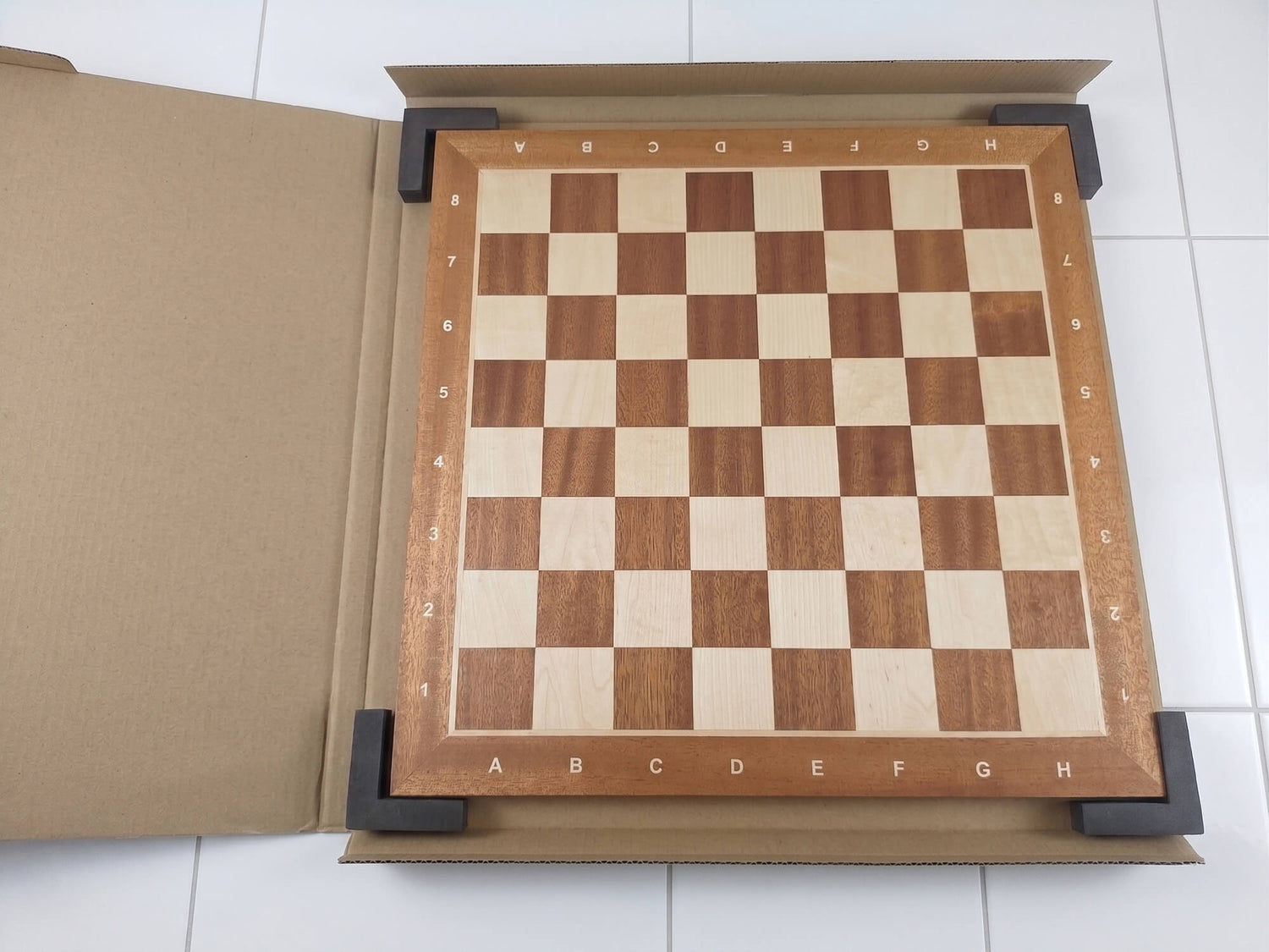 The Monarch Series mahogany and maple chess board secured in its protective cardboard packaging box with foam corner guards, against a white tiled floor.