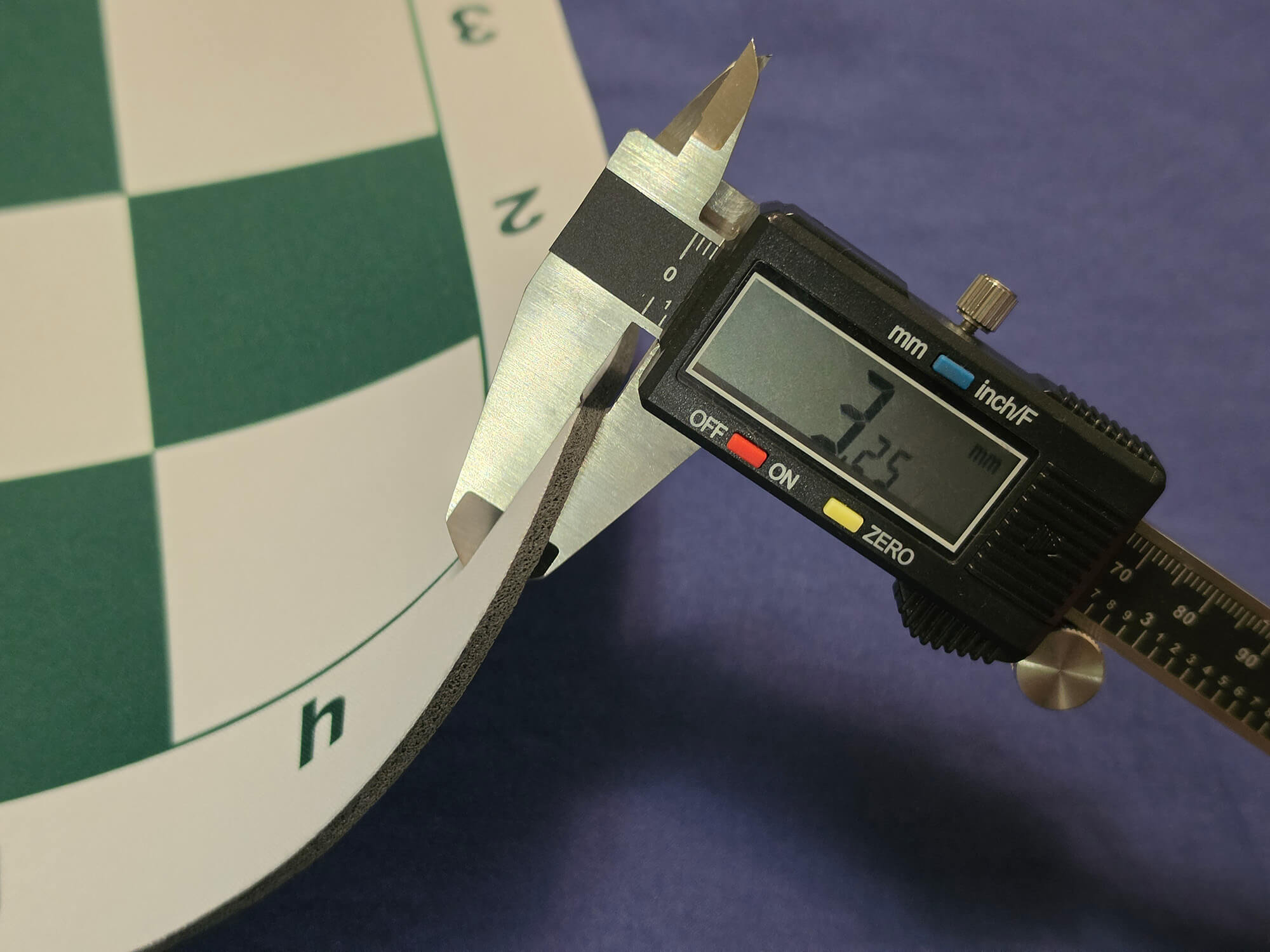 Close-up of a digital micrometer caliper measuring the thick, cushion thickness of a green and white mousepad chess board.