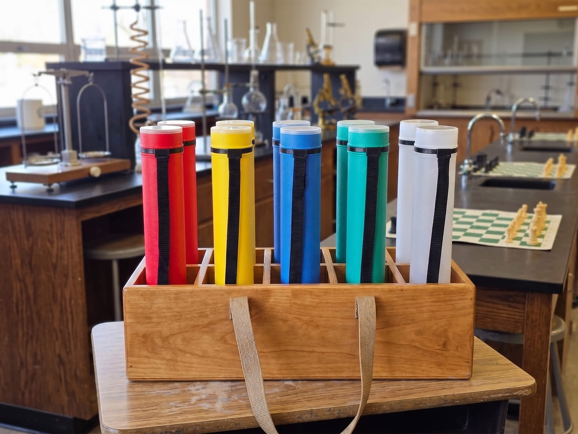 A group of colorful Sentry telescopic chess tubes in red, yellow, blue, green, and white organized in a solid wood carrying case with fabric handles, set against a classroom laboratory background with an unset chess game.