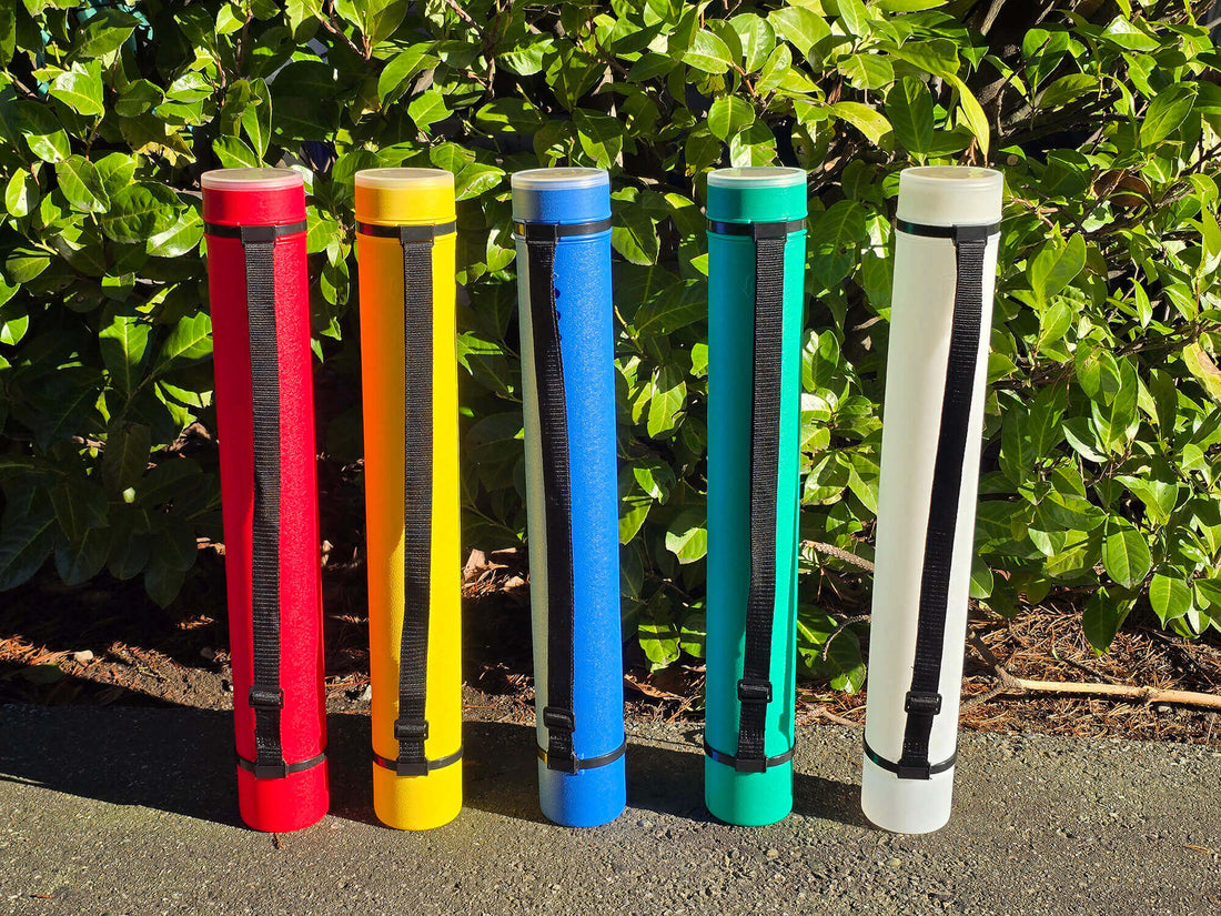 Five plastic chess storage tubes, in colours red, yellow, blue, green, and white, lined up side-by-side on a concrete surface in front of a green leafy hedge under bright sunlight. Each tube has a black adjustable shoulder strap and a clear lid.