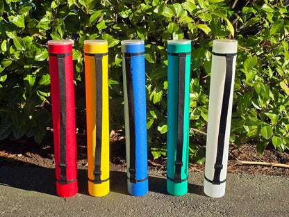 Five plastic chess storage tubes, in colours red, yellow, blue, green, and white, lined up side-by-side on a concrete surface in front of a green leafy hedge under bright sunlight. Each tube has a black adjustable shoulder strap and a clear lid.