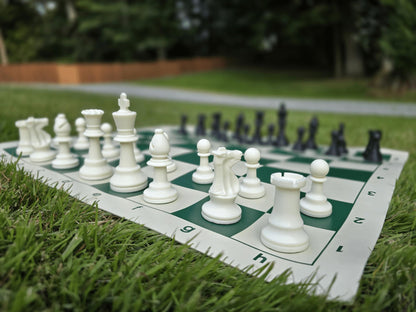 Standard plastic Staunton chess pieces on a green and white roll-up vinyl board, shown in a natural outdoor setting on green grass.