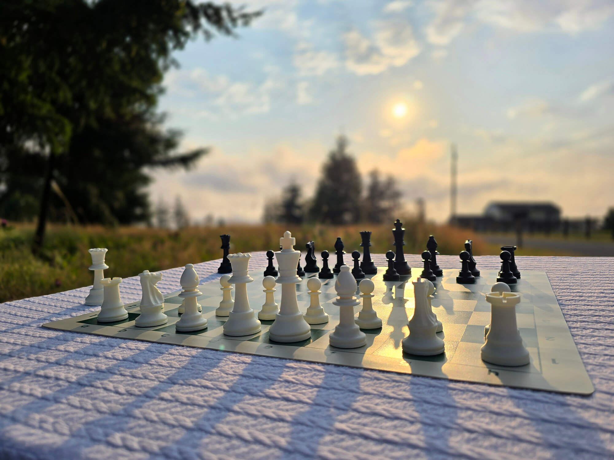 Tournament Chess Set on a Vinyl Chess Board on a White, Knitted Tablecloth in a Meadow with a Setting Sun, and Blurred Background with a Farmhouse in the Distance