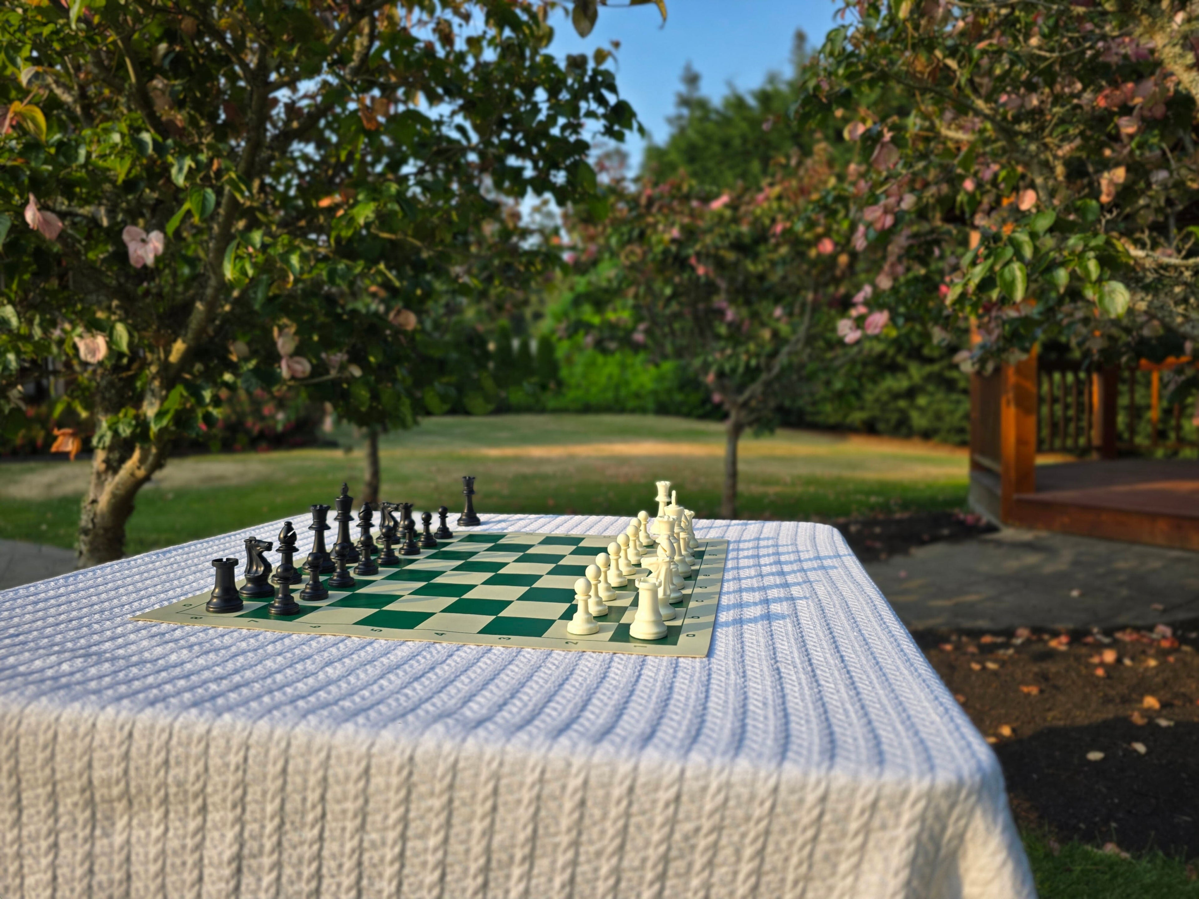 Tournament Chess Set on a Vinyl Chess Board on a White, Knitted Tablecloth in a Garden with a Gazebo and Trees with Pink Flowers