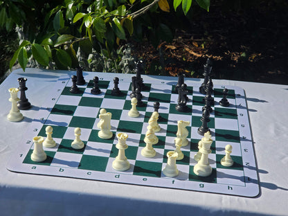 A complete tournament chess set with plastic pieces set up on a green and white mousepad board, captured in bright outdoor sunlight.