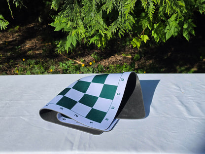 Green and white mousepad chess board with algebraic notation, shown folded twice on a white tabletop outdoors to demonstrate its flexible, wrinkle-resistant rubber backing.